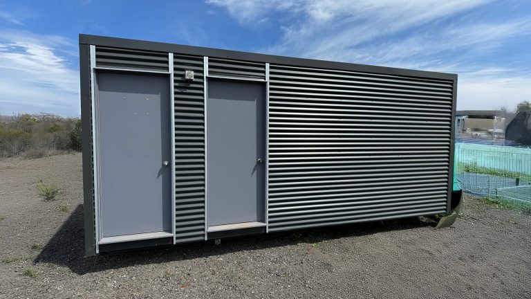 Grey corrugated metal site shed with dual doors on gravel lot.