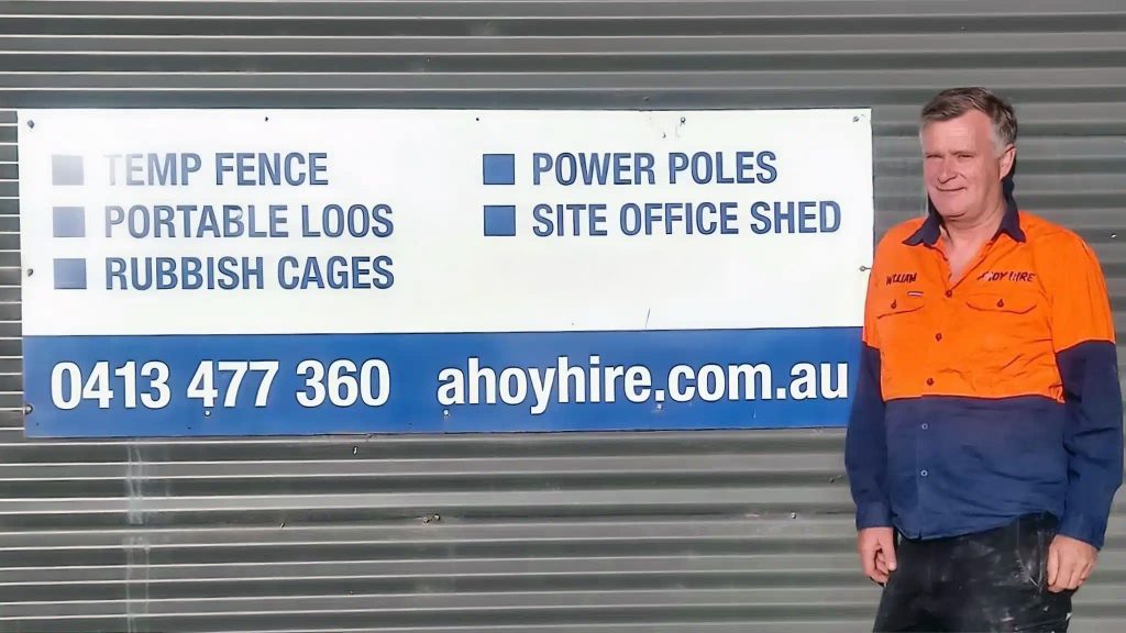 Will standing next to a company sign listing Melbourne construction services including temp fence, portable loos, rubbish cages, power poles, and site office sheds.